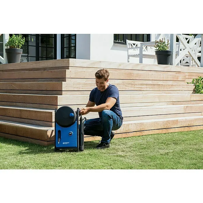 Nilfisk Hidrolimpiadora de alta presión CORE 150 PC - 150 bar, 2.000 W, Capacidad de bombeo: 468 l/h - Un hombre está preparando una limpiadora de alta presión azul Nilfisk con carrete de manguera frente a una terraza de madera al aire libre.