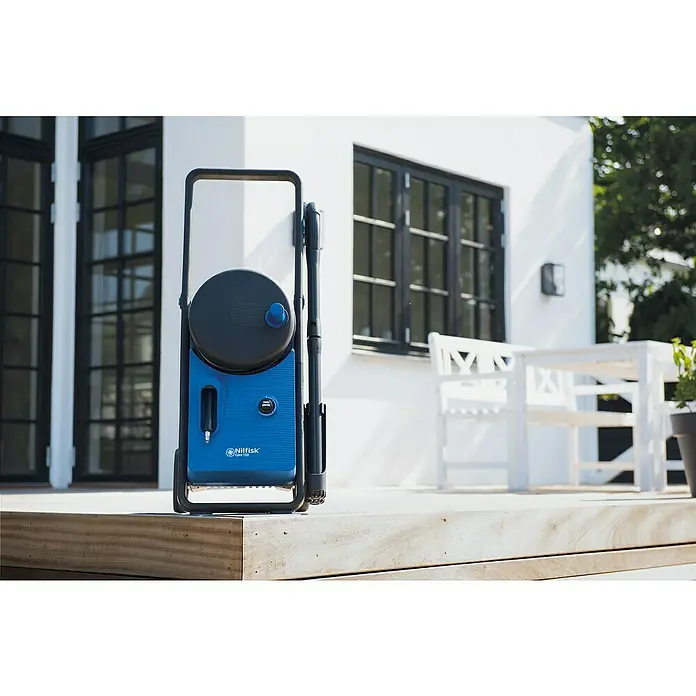 Nilfisk Hidrolimpiadora de alta presión CORE 150 PC - 150 bar, 2.000 W, Capacidad de bombeo: 468 l/h - Primer plano de una hidrolimpiadora azul Nilfisk Core 150 en una terraza de madera frente a una casa moderna con paredes blancas y ventanas negras.