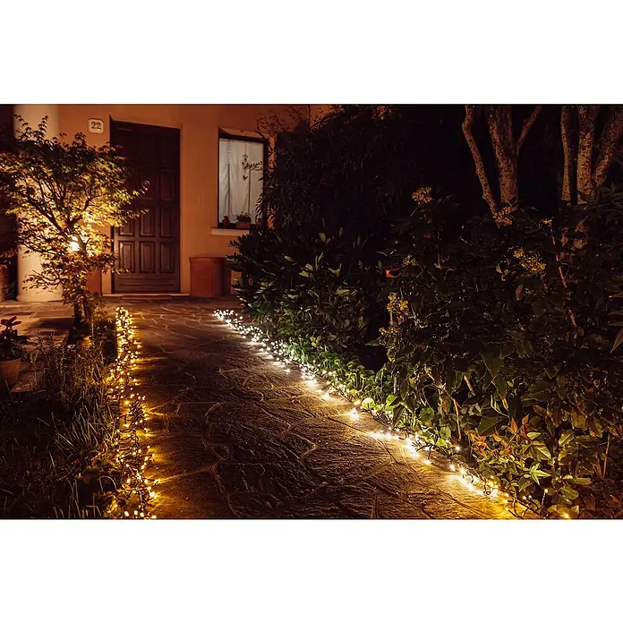 Twinkly Cadena de luces LED Cluster - Para exterior, Largo: 6 m, 400 ud., Funcionamiento en red, Color de luz: Ámbar - Camino iluminado con luces que conduce a una casa con puerta y ventanas de madera, rodeado de exuberante vegetación.