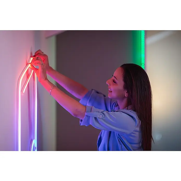 Twinkly Tira de luces LED Flex - Largo: 2 m, Color de luz: RGB, 15 W - Mujer colocando una decoración de neón vertical brillante en una pared, creando un ambiente moderno y colorido.
