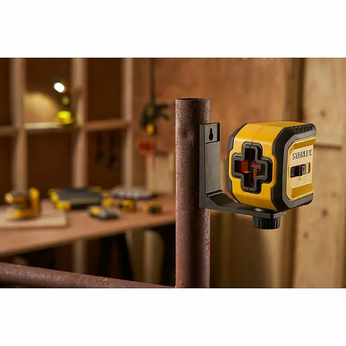 Stanley Nivel láser en cruz STHT77611-0 - Zona de trabajo máx.: 10 m - Primer plano de un láser de líneas cruzadas Stanley amarillo conectado a un riel de metal con herramientas al fondo.