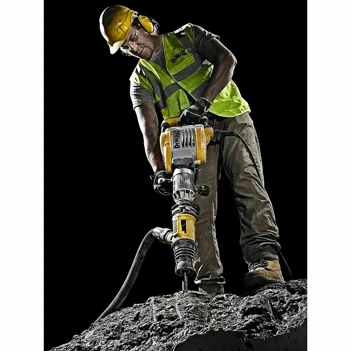 DeWalt Martillo perforador D25902K - 1.550 W, 19 J, Alojamiento SDS-MAX - Un trabajador de la construcción utiliza un martillo neumático DeWalt para perforar la piedra. El equipo de protección incluye casco, gafas y protección auditiva.