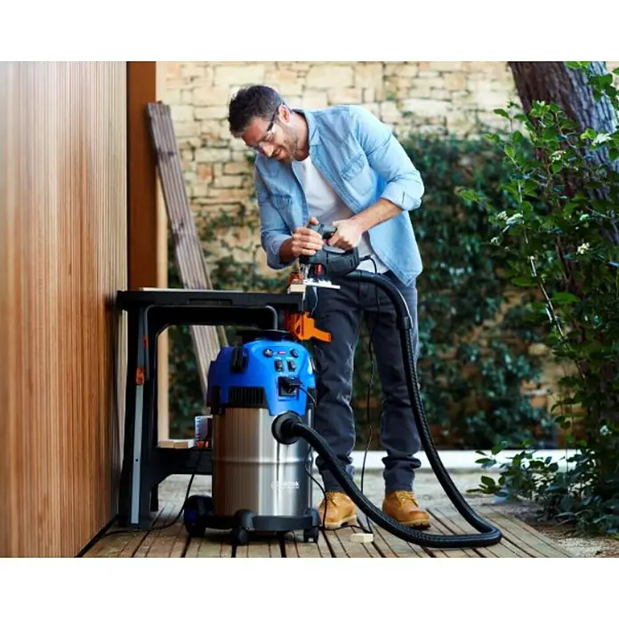 Nilfisk Elektro-Nass- & Trockensauger Multi II 22 Inox - 1.200 W, Behältervolumen: 22 l, Mit Beutel - Ein Mann benutzt einen Nilfisk Nass- und Trockensauger beim Sägen von Holz im Freien. Der Sauger ist blau und silber.
