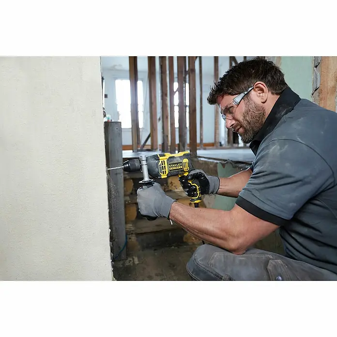 Stanley FatMax Schlagbohrmaschine FMEH750K-QS - 750 W, Schnellspannbohrfutter - Ein Mann bohrt mit einer gelben Stanley-Bohrmaschine in eine Wand, Schutzbrille und Handschuhe schützend.