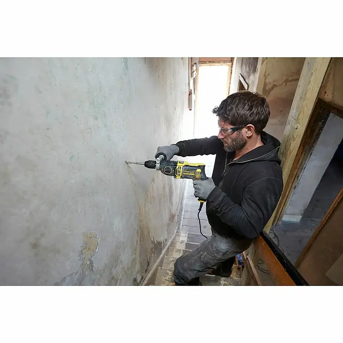 Stanley FatMax Schlagbohrmaschine FMEH1100K-QS - 1.100 W, Schnellspannbohrfutter - Ein Mann bohrt mit einer Stanley Fatmax Schlagbohrmaschine ein Loch in eine Wand. Der Mann trägt dabei eine Schutzbrille und Handschuhe.