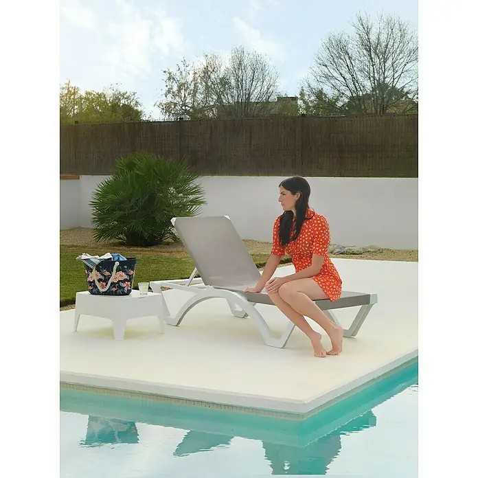 Tumbona de jardín Mykonos - 74 x 190 x 36 cm, Arena, Respaldo regulable - Una mujer está sentada en una tumbona gris junto a la piscina con una bolsa y una bebida en una mesa cercana. Está disfrutando del sol en un día de verano.