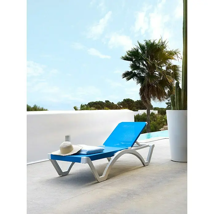 Tumbona de jardín Mykonos - 74 x 190 x 36 cm, Azul, Plástico, Respaldo regulable - Una tumbona azul con un sombrero y una toalla se sitúa en un patio con vistas a una piscina y palmeras. El lugar perfecto para relajarse.