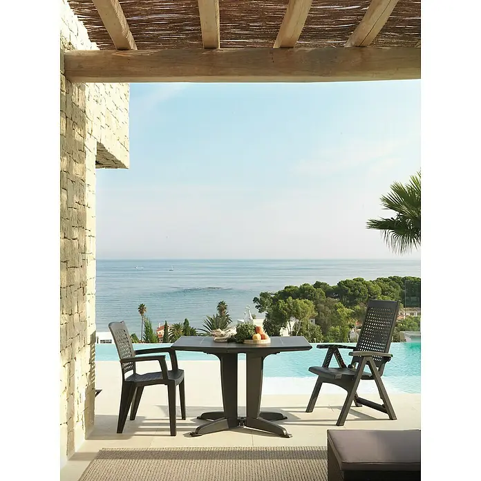Silla de jardín Atlantic - 59 x 60 x 87 cm, Antracita, Polipropileno, Apilable - Una mesa de jardín con dos sillas está ubicada en una terraza con vistas al mar. La mesa está decorada con una botella y fruta.