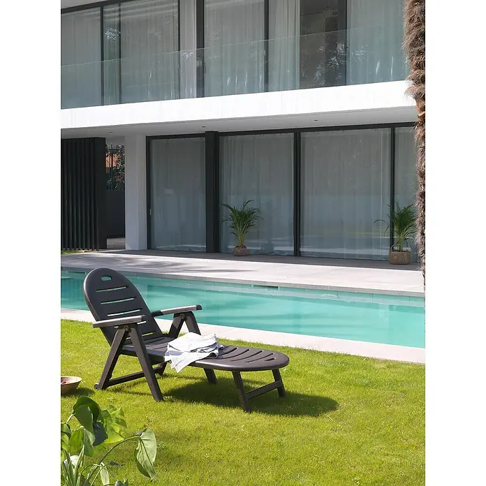 Tumbona de jardín Caimán - 72 x 195 x 100 cm, Wengué, Plástico, Respaldo regulable - Una tumbona gris con una toalla está en un césped verde frente a una casa moderna con piscina y grandes ventanales.