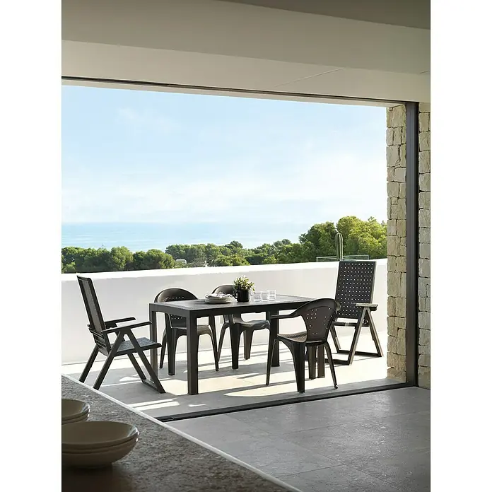 Mesa de jardín Dream - L x An x Al: 140 x 85 x 73 cm, Polipropileno, Wengué - Conjunto de comedor exterior de plástico negro en una terraza con vistas al mar. Incluye mesa, sillas y tumbona.