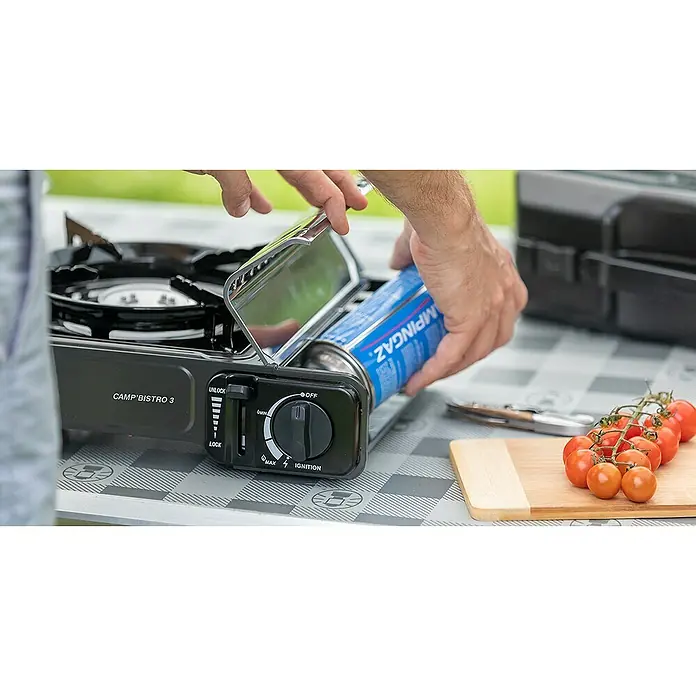 Campingaz Hornillo de gas Bistro 3 - L x An x Al: 36 x 32 x 13,7 cm, 2,2 W, Número de quemadores: 1 ud. - Primer plano de un hornillo de gas Camp'Bistro 3 al que se le está cargando un cartucho de gas azul, junto a tomates y un cuchillo sobre una mesa.