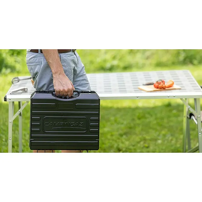 Campingaz Hornillo de gas Bistro 3 - L x An x Al: 36 x 32 x 13,7 cm, 2,2 W, Número de quemadores: 1 ud. - Persona sosteniendo una funda negra de Campingaz frente a una mesa con comida.
