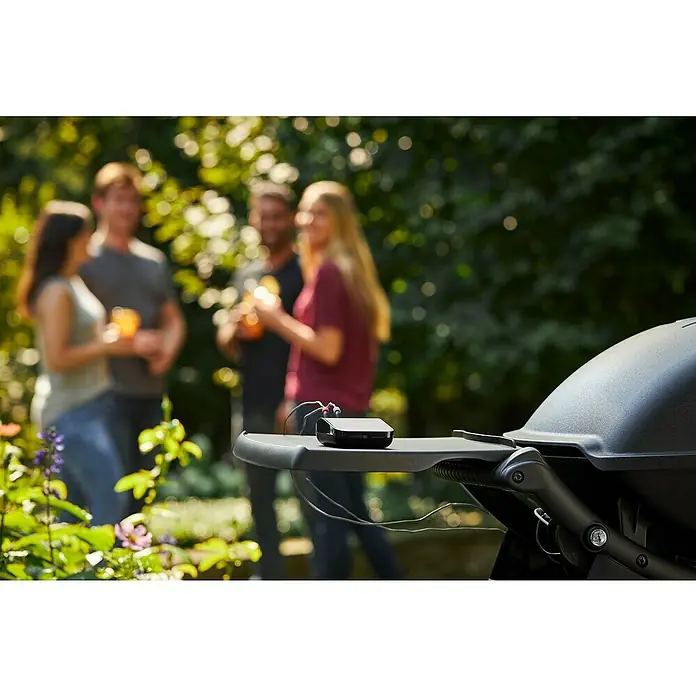 Weber Barbacoa de gas Q2200 - Superficie de la parrilla (An x Pr): 54 x 39 cm, 3,52 kW, Número de quemadores: 1 - Toma de una parrilla en un entorno de patio trasero, que muestra un dispositivo negro sentado en la mesa lateral de la parrilla. Hay gente en el fondo.