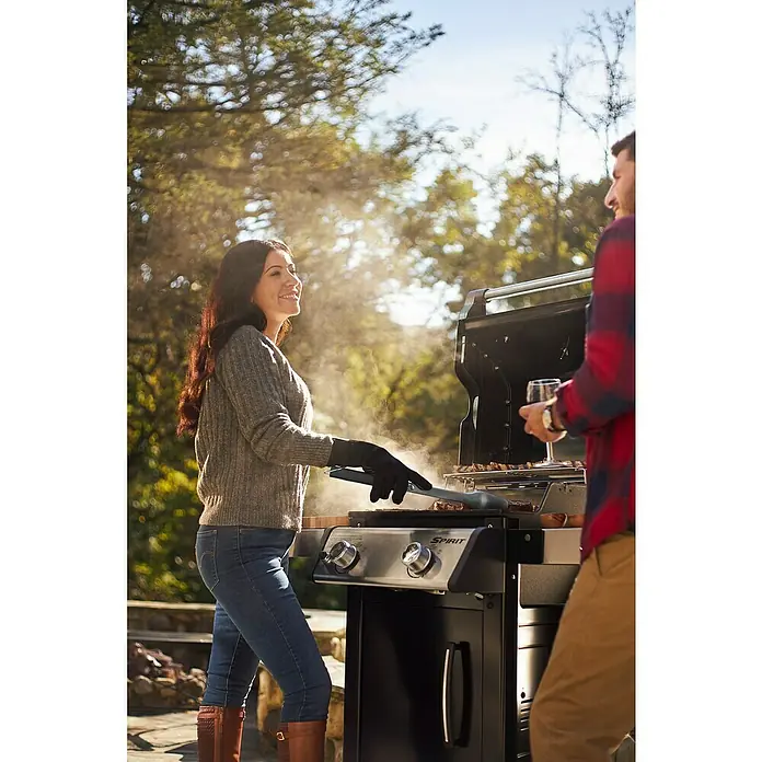 Weber Barbacoa de gas Spirit E-215 GBS - Superficie de la parrilla (An x Pr): 52 x 45 cm, Número de quemadores: 2 - Una pareja haciendo una parrillada al aire libre en una parrilla de gas Spirit, la mujer usando guantes y pinzas para asar. Árboles y algo de sol son visibles al fondo.