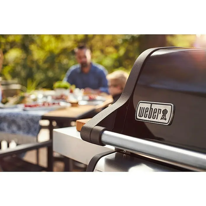 Weber Barbacoa de gas Spirit E-215 GBS - Superficie de la parrilla (An x Pr): 52 x 45 cm, Número de quemadores: 2 - Primer plano de una parrilla Weber al aire libre, con la familia al fondo. Asar con Weber.
