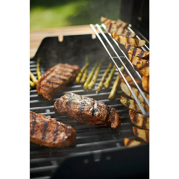Weber Barbacoa de gas Spirit E-215 GBS - Superficie de la parrilla (An x Pr): 52 x 45 cm, Número de quemadores: 2 - Primer plano de bistecs, espárragos y gajos de patata cocinándose en una parrilla con deliciosas marcas de chamuscado.