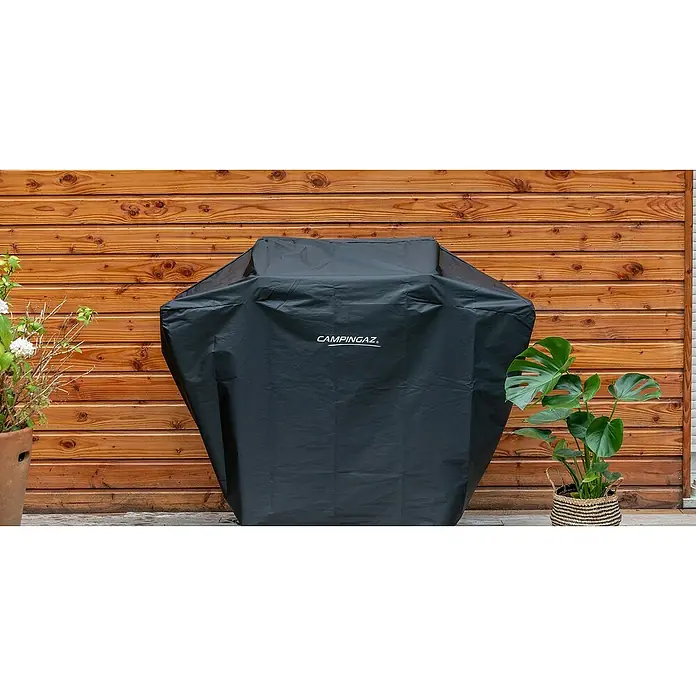 Campingaz Funda protectora para barbacoa Universal - L x An x Al: 146 x 118 x 66 cm, Poliéster, Negro - La imagen muestra una cubierta de barbacoa Campingaz frente a una pared de madera, con plantas en macetas a izquierda y derecha.