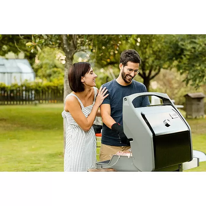 Weber Barbacoa de gas Spirit II E-310 + 7 accesorios - Superficie de la parrilla (An x Pr): 60 x 46 cm, 8,7 kW, Número de quemadores: 3 - Un hombre con guante de barbacoa negro cocina en una parrilla negra y plateada, con una mujer a su lado.