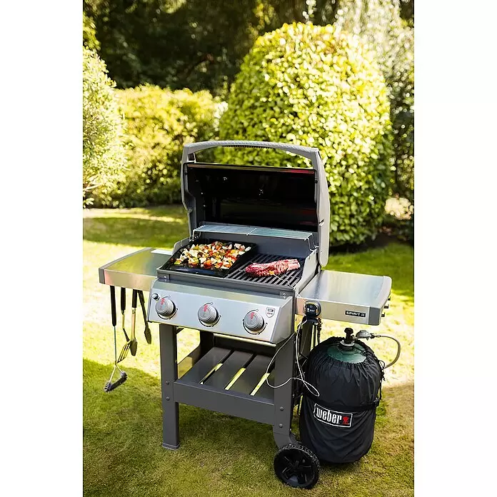 Weber Barbacoa de gas Spirit II E-310 + 7 accesorios - Superficie de la parrilla (An x Pr): 60 x 46 cm, 8,7 kW, Número de quemadores: 3 - Una barbacoa de gas Weber Spirit II gris y plateada en el exterior, asando verduras y un filete. Un tanque de propano con funda negra Weber está conectado.
