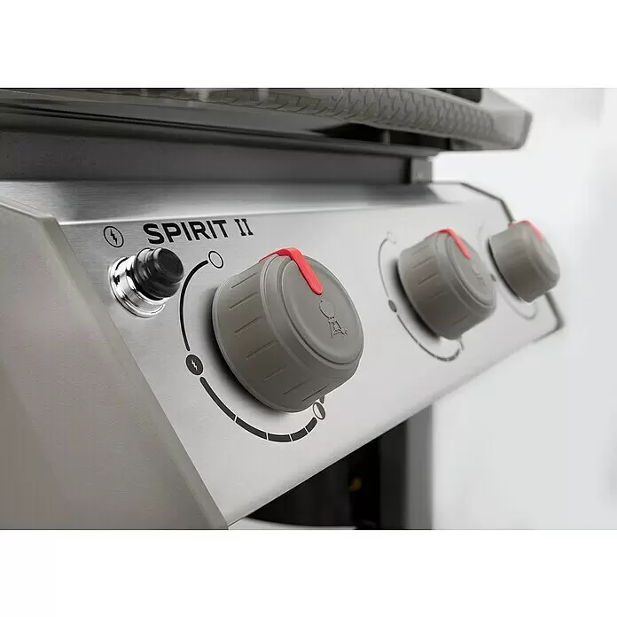 Weber Barbacoa de gas Spirit II E-310 + 7 accesorios - Superficie de la parrilla (An x Pr): 60 x 46 cm, 8,7 kW, Número de quemadores: 3 - Primer plano del panel de control de una parrilla de acero inoxidable cepillado con botón de encendido negro y tres perillas grises con indicadores rojos. 'SPIRIT II' impreso.