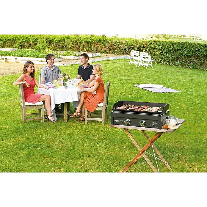 Campingaz Barbacoa de gas L - Superficie de la parrilla (An x Pr): 66 x 42 cm, Número de quemadores: 2 - La imagen muestra una parrilla Campingaz Plancha L sobre una mesa de madera mientras la gente cena en un jardín.