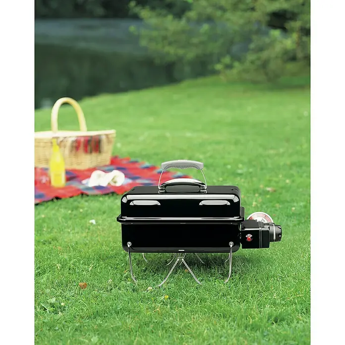 Weber Go-Anywhere Holzkohlegrill - Grillfläche (B x T): 42 x 26 cm, Schwarz - Tragbarer schwarzer Grill mit Picknickkorb im Hintergrund, ideal für ein entspanntes Essen im Freien.