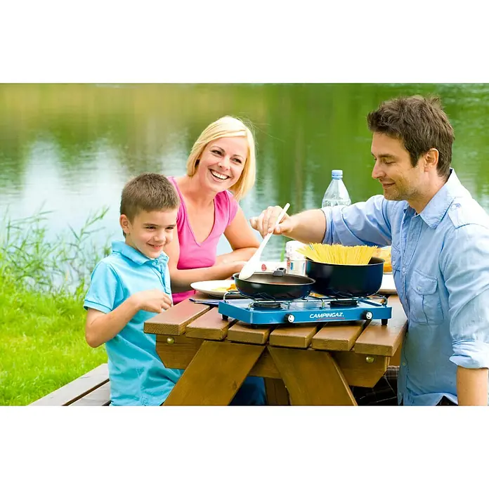 Campingaz Hornillo de gas Base Camp Lid - 43 x 21 x 8,5 cm, 1,6 W - Una cocina de gas Campingaz azul sobre una mesa de madera mientras una familia cocina al aire libre. ¡Ideal para cocinar en todas partes!