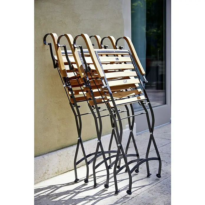 Sunfun Gartenmöbel-Set Diana/Moni - 5 -tlg., Holz, Naturbraun/Schwarz, Tischplatte ausziehbar - Stapel aus klassischen Klappstühlen aus Holz und Metall, die an einer Wand im Freien lehnen und Schatten auf den Boden werfen.