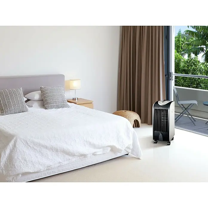 Purline Rafy Climatizador evaporativo 51 - Blanco/Negro, Altura: 58 cm, 75 W - Un enfriador de aire portátil se encuentra en un dormitorio luminoso cerca de una ventana de balcón, prometiendo alivio fresco en días calurosos.