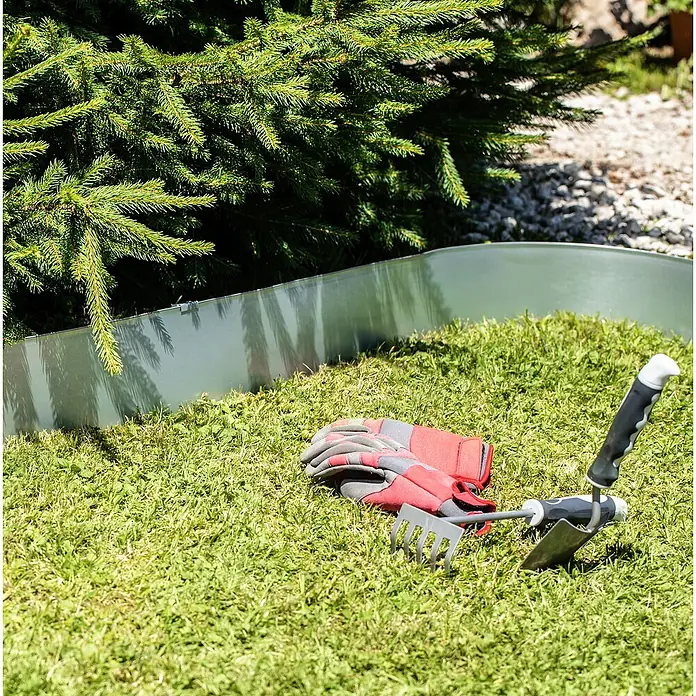 Windhager Rasenkante - 15 cm x 5 m, Befestigung der Rasenkanten, Material: Metall - Gartenarbeitsszene mit Rasenkante, Handschuhen, Harke und Schaufel unter einem grünen Nadelbaum, die zum Gärtnern einlädt.