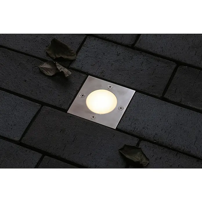 Paulmann Plug & Shine Bodeneinbauleuchte - 3,6 W, Warmweiß, IP65, Quadratisch - Nahaufnahme einer quadratischen Bodeneinbauleuchte mit warmem Licht, eingebettet in dunkles Pflaster und umgeben von trockenem Laub.