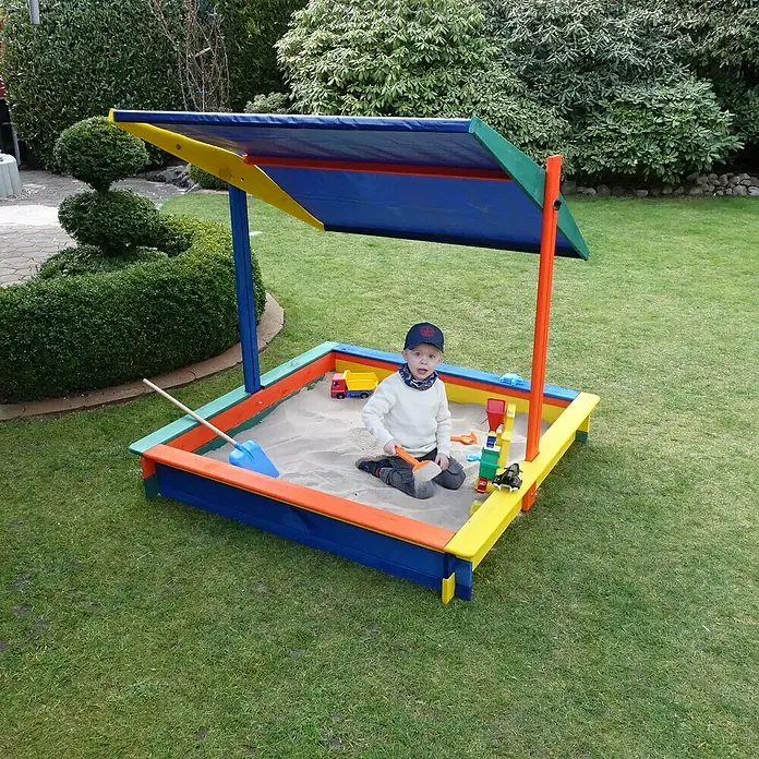 Promadino Sandkasten Nadine140 x 140 x 13 cm, Kiefer, Höhenverstellbares Dach - Ein Junge spielt im Sandkasten mit Sonnenschutz, der in fröhlichen Farben gehalten ist, und hat sichtlich Spaß dabei.