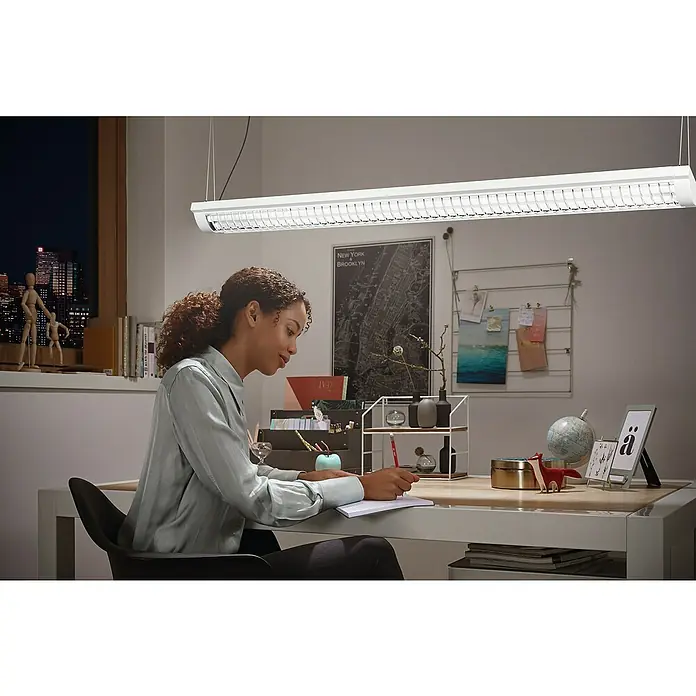 Ledvance LED-Deckenleuchte Office Line - 25 W, L x B x H: 59,5 x 13,4 x 4,6 cm, Weiß, Kaltweiß - Eine Frau sitzt an einem Schreibtisch unter einer modernen, länglichen Büroleuchte, die helles Licht spendet und die Arbeitsfläche optimal ausleuchtet.