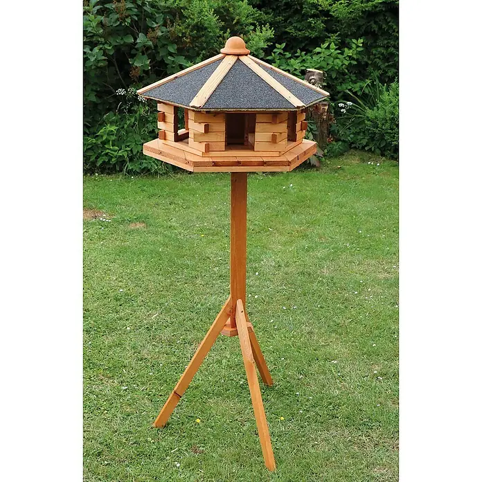 Promadino Vogelhaus Fehmarn - Holz, Hellbraun, Ausstattung: Vogelhausständer, Mit Silo - Ein hölzernes Vogelhaus auf einem Ständer im Garten. Das Vogelhaus hat ein dunkles Dach und steht auf einer gepflegten Rasenfläche.