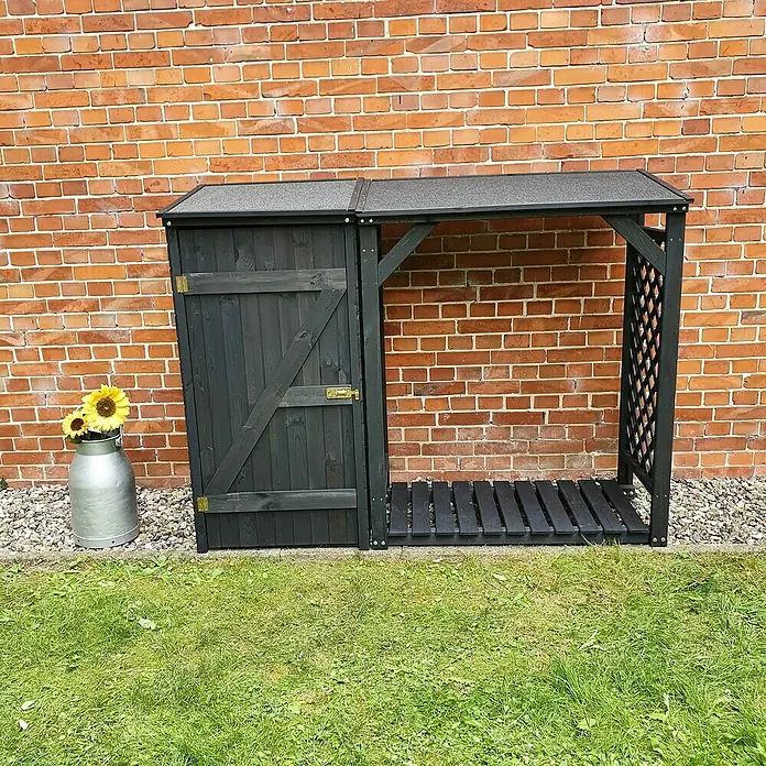 Promadino Kaminholzregal Kombi Speyer - 192 x 69 x 148 cm, Anthrazit - Ein dunkelgrauer Holzschuppen mit Gitterstruktur steht vor einer roten Backsteinmauer, daneben ein Metallgefäß mit Sonnenblumen.