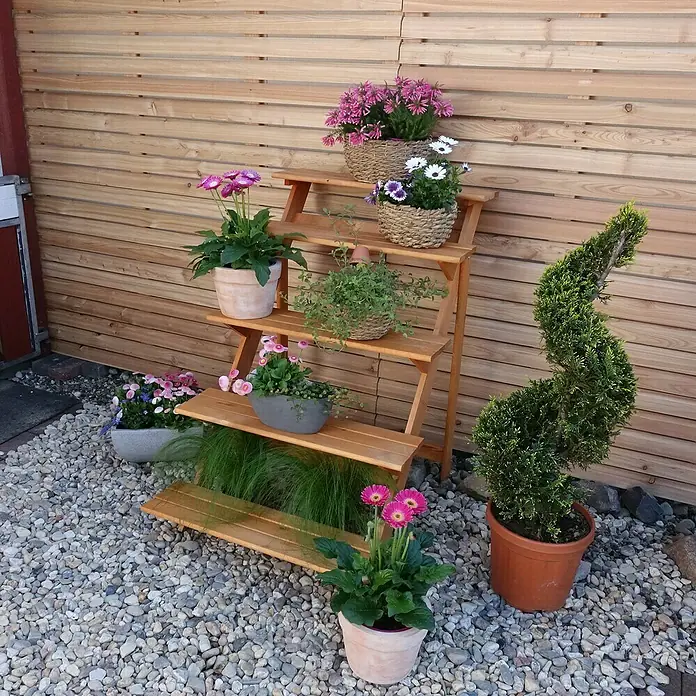 Promadino Blumentreppe - 100 x 78 x 109 cm, Kiefer, Braun - Blumenregal aus Holz mit bunten Blumen in Töpfen und Körben auf einer Terrasse mit Kiesboden vor einer Holzwand.