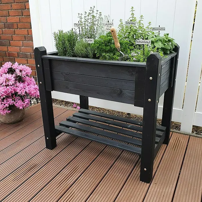 Promadino Kräuterhochbeet - L x B x H: 90 x 60 x 84,5 cm, Anthrazit - Hochbeet aus schwarzem Holz mit Kräutern bepflanzt, auf einer Holzterrasse platziert, daneben ein Blumentopf mit rosa Blumen.
