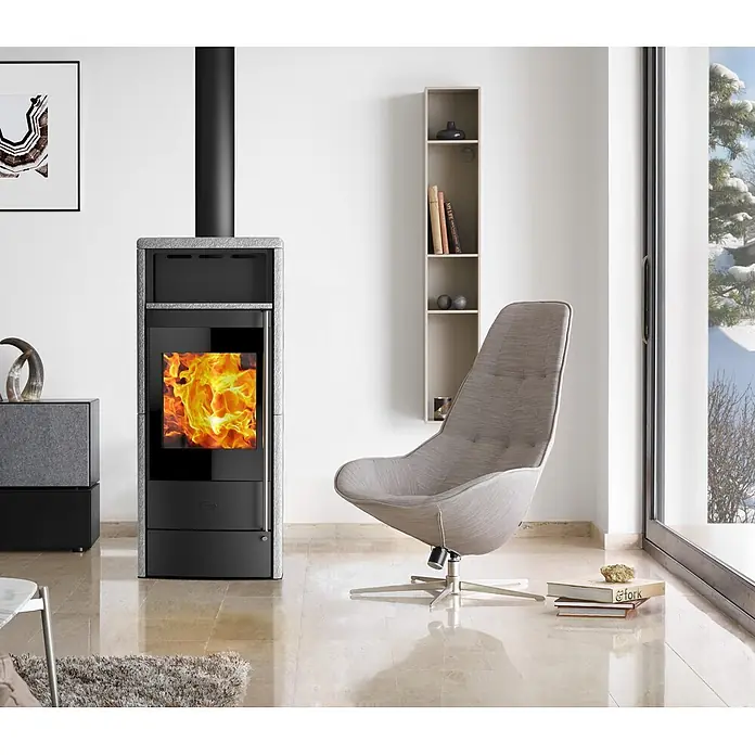 Fireplace Kaminofen Teramo RLU - 6,4 kW, Raumheizvermögen: 108 m³, Material Verkleidung: Speckstein, Schwarz - Ein moderner Kaminofen mit loderndem Feuer und ein stilvoller Sessel schaffen eine gemütliche Atmosphäre im Wohnzimmer.