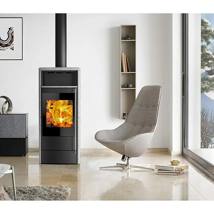 Fireplace Kaminofen Teramo RLU - 6 kW, Raumheizvermögen: 108 m³, Material Verkleidung: Speckstein, Schwarz - Ein moderner Kaminofen mit loderndem Feuer und ein stilvoller Sessel schaffen eine gemütliche Atmosphäre im Wohnzimmer.