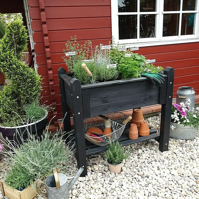 Promadino Kräuterhochbeet - L x B x H: 90 x 60 x 84,5 cm, Anthrazit - Ein erhöhter Kräutergarten aus schwarzem Holz mit verschiedenen Kräutern, Töpfen und Gartenwerkzeugen auf einem Kiesweg vor einem roten Haus.