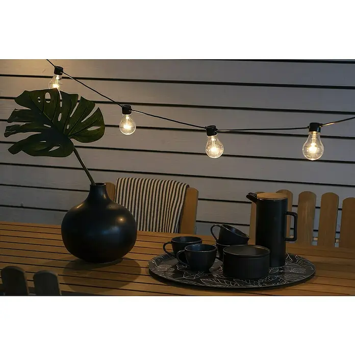 Konstsmide LED-Lichterkette S - Außen, Lichtfarbe: Bernstein - Tischdekoration mit einer schwarzen Vase, Teegeschirr und Lichterkette, die eine gemütliche Atmosphäre für den Außenbereich schafft.