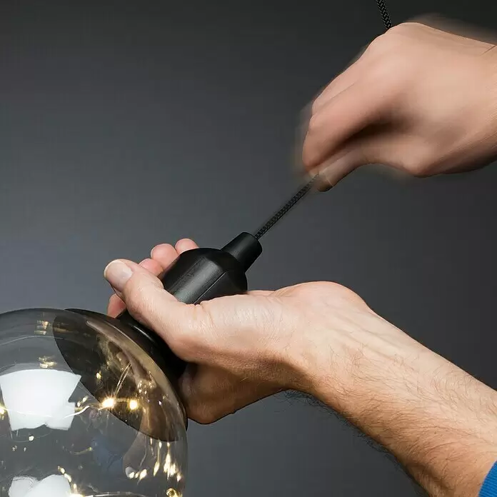 Konstsmide LED-Dekoleuchte - 0,6 W - Hände montieren eine schwarze Lampenfassung an eine große klare Glühbirne mit integrierten LED-Lichterketten und ziehen am geflochtenen Kabel.
