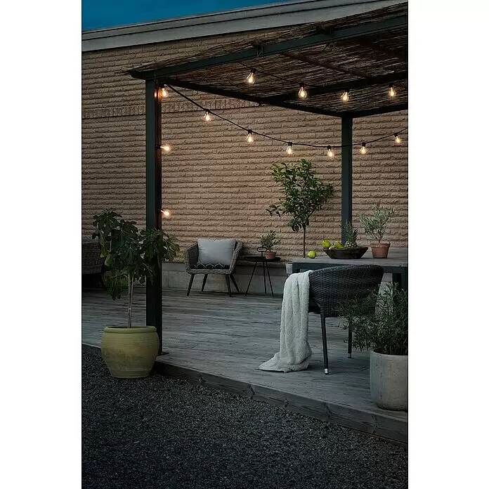 Konstsmide LED-Lichterkette - Außen, Lichtfarbe: Bernstein - Außenbereich mit Pergola, beleuchtet von warmen Lichterketten, Gartenmöbeln und Topfpflanzen.