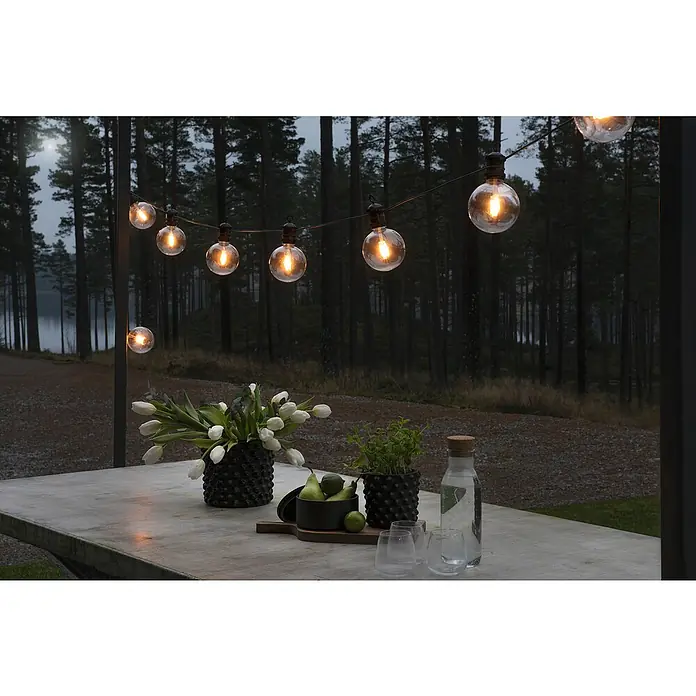 Konstsmide LED-Lichterkette System Erweiterung - Außen, Lichtfarbe: Bernstein - Eine Lichterkette mit runden Glühbirnen hängt über einem Tisch mit Blumen, Obst und Getränken, die eine gemütliche Atmosphäre schafft.