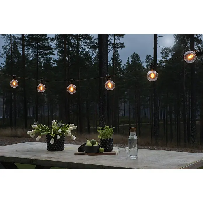 Konstsmide LED-Lichterkette Startset - Außen, 10 m, Lichtfarbe: Bernstein - Festliche Szene im Freien mit einer Lichterkette, die eine einladende Atmosphäre über einem gedeckten Tisch mit Blumen und Getränken schafft.