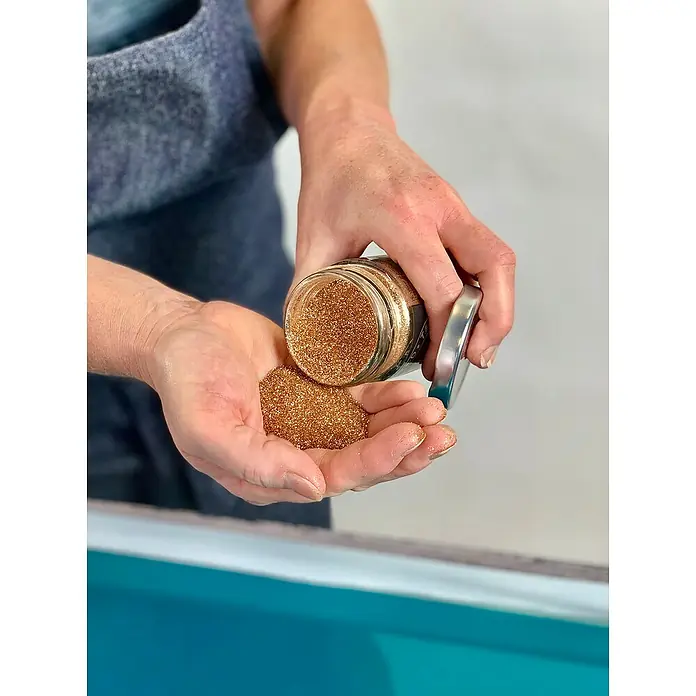 Fleur Purpurina Glitter - Dorado, 90 g, Brillante - Una persona está vertiendo purpurina dorada de un frasco en su mano, centrándose en el material de manualidades brillante.
