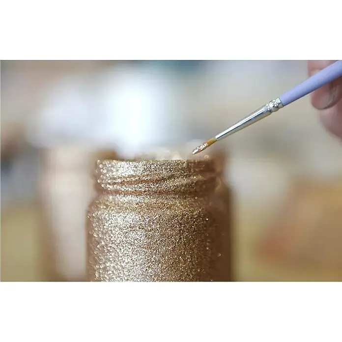 Fleur Purpurina Glitter - Champagne, 90 g, Brillante - Primer plano de un pincel tomando pintura dorada con purpurina de un pequeño frasco de vidrio, ideal para proyectos artísticos y decoraciones.