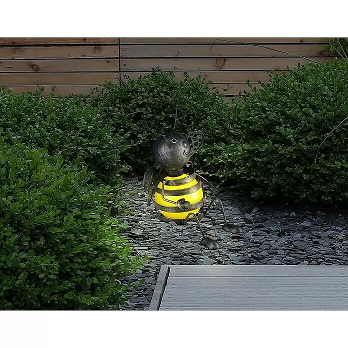 Globo Solar-Dekoleuchte Biene - 0,06 W, L x B x H: 210 x 190 x 240 mm, Schwarz, Gelb, IP44 - Eine dekorative Bienen-Solarleuchte aus Metall, gelb-schwarz gestreift, leuchtet sanft zwischen grünen Gartenbüschen und dunklem Kies.