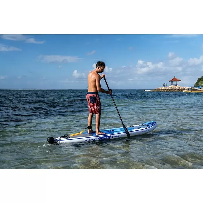 Sublue Halterung Tini SUP Paddleboard Set - Passend für: Sublue Tauchscooter Tini - Ein Mann steht auf einem blau-weißen Paddleboard in klarem Flachwasser und paddelt, mit Meereshintergrund.