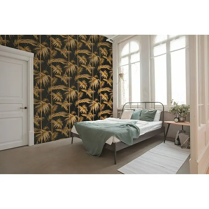A.S. Création Metropolitan Stories Vliestapete Gräser - Schwarz/Gold, Floral, 10,05 x 0,53 m - Ein Schlafzimmer mit einer schwarzen Tapete mit goldenen Palmen, einem Bett mit grünen Decken und einem Holztisch.